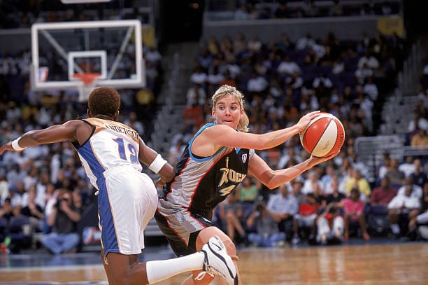 Suzie McConnell-Serio holding a basketball in a basketball game, with an opposing player running past her. McConnell-Serio looking ahead in black jersey with silver sides of jersey. 
